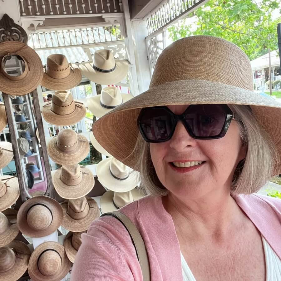 Woman wearing a wide-brimmed hat and sunglasses smiles for a selfie in front of a display of Tula Hats and other straw hats at an outdoor market.
