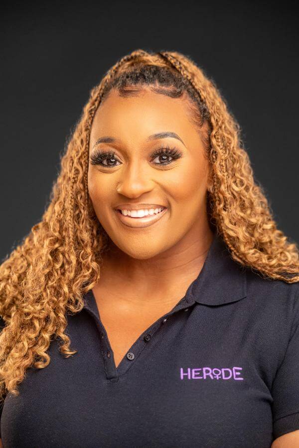 A Ride-Sharing App Driven by Women? We Love it! - 3 A woman with long curly hair smiles at the camera, wearing a dark polo shirt proudly featuring the HERIDE logo embroidered in purple.