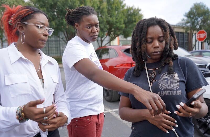 A Ride-Sharing App Driven by Women? We Love it! - 4 Three people stand in a parking lot; one holds a phone displaying the Heride app while another points at its screen, and the third looks on. Cars and a stop sign are visible in the background.