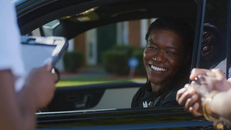A Ride-Sharing App Driven by Women? We Love it! - 1 A smiling person sits in the driver's seat of a heride car while interacting with two people outside the vehicle, one holding a clipboard.