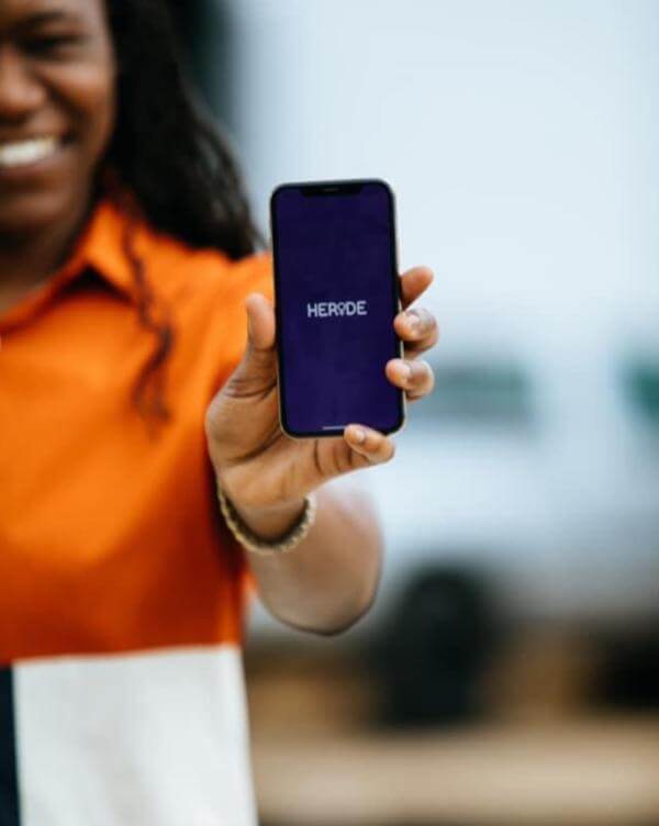 A Ride-Sharing App Driven by Women? We Love it! - 2 A person holds a smartphone displaying the word "HERODE" on its screen, while wearing an orange, white, and blue shirt—perhaps checking out the latest offer from Heride.