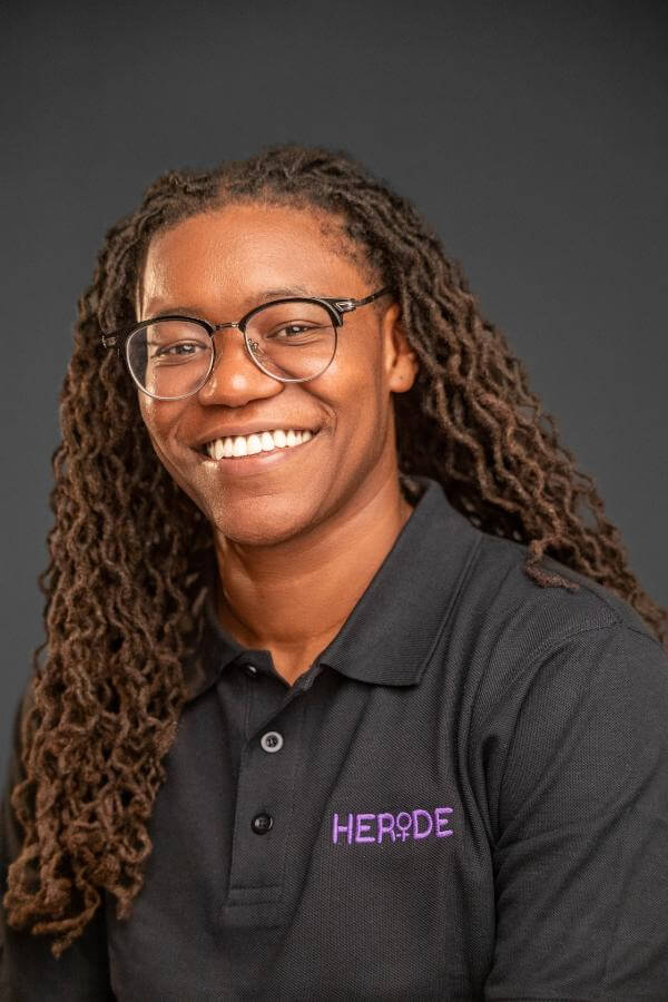 A Ride-Sharing App Driven by Women? We Love it! - 5 Person with long, braided hair and glasses smiles at the camera, wearing a black collared shirt with the purple heride logo on a plain dark background.