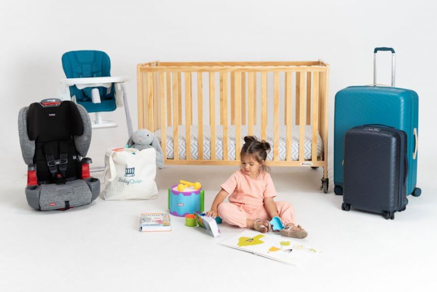 A young child sits on the floor reading a book, surrounded by a crib, high chair, car seat, suitcases, a bag, toys, children's books, and other babyquip essentials—all set against a plain white background.