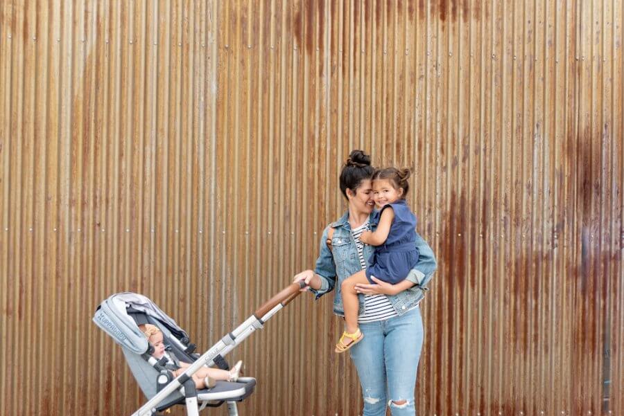 A woman in jeans holds a young girl on her hip and stands next to a stroller with a baby in it, rented from BabyQuip, in front of a corrugated metal wall.