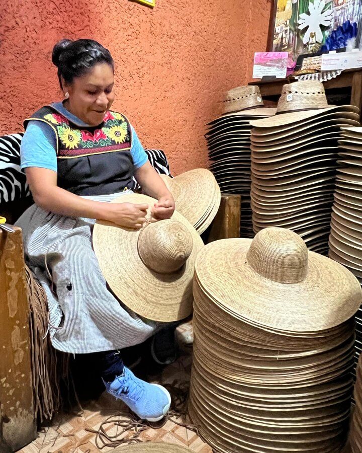 A woman sits indoors weaving a large straw Tula Hat by hand, surrounded by stacks of finished Tula Hats against an orange wall.