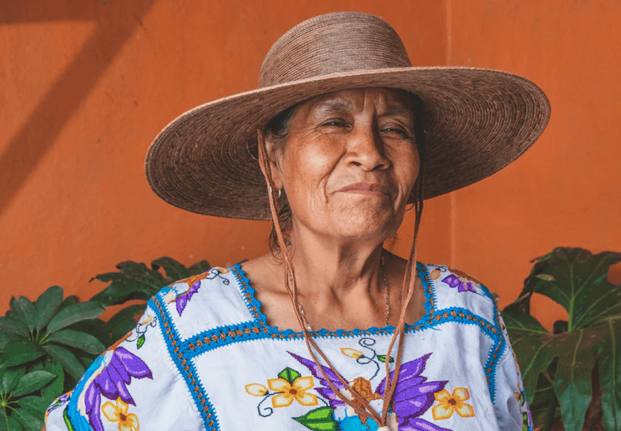 Older woman wearing a Tula Hat and a white blouse with colorful floral embroidery stands in front of an orange wall and green plants.