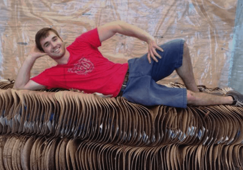 A man in a red t-shirt and blue shorts, sporting a stylish Tula Hat, poses lounging on a stack of flattened cardboard boxes in front of a plastic-covered wall.