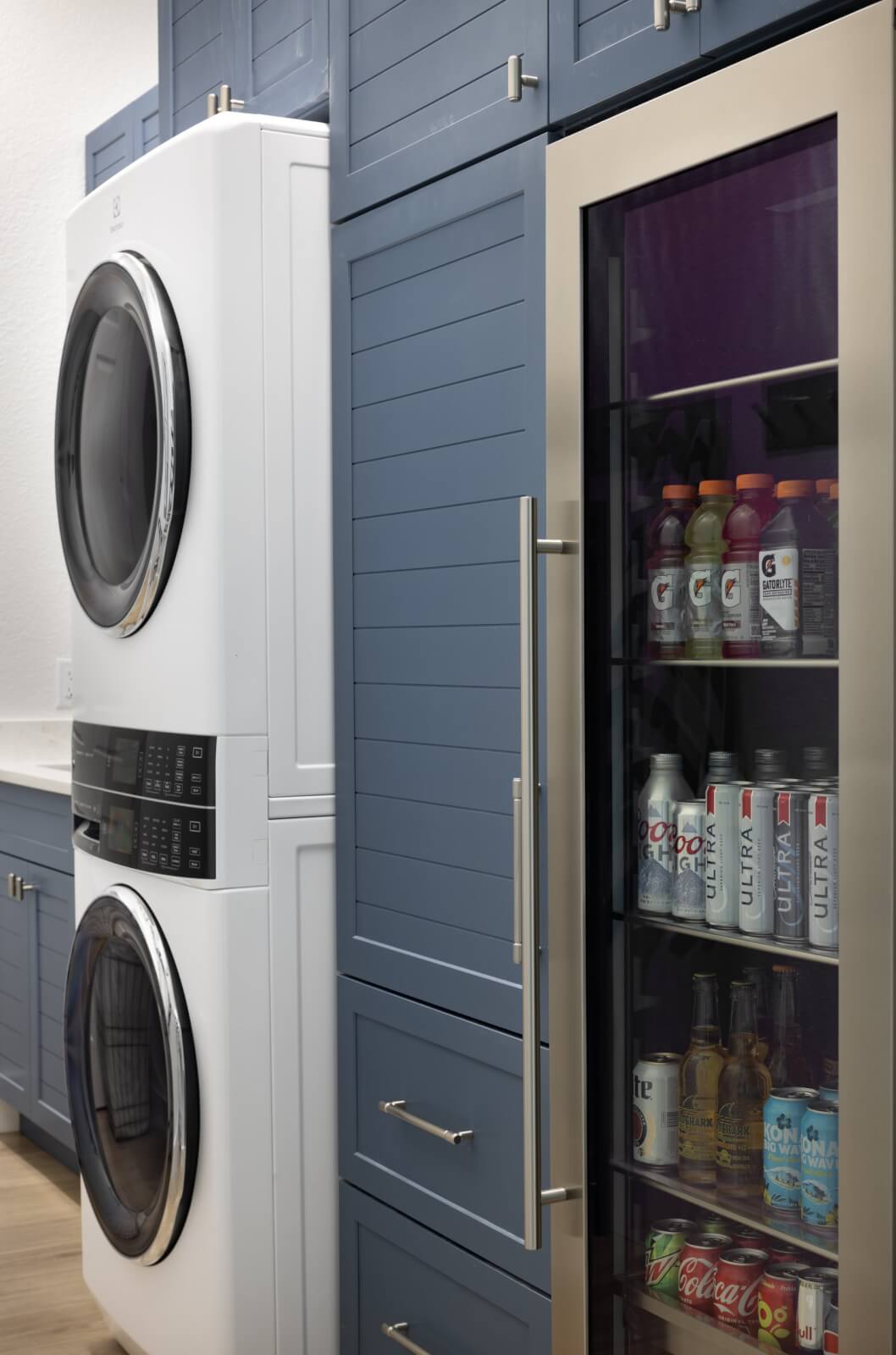 Look Inside This Effortlessly Chic Cape Haze Fishing Retreat - 13 A laundry room with stacked washer and dryer next to a glass-front beverage fridge containing sodas, sports drinks, and bottled water, surrounded by blue cabinetry.