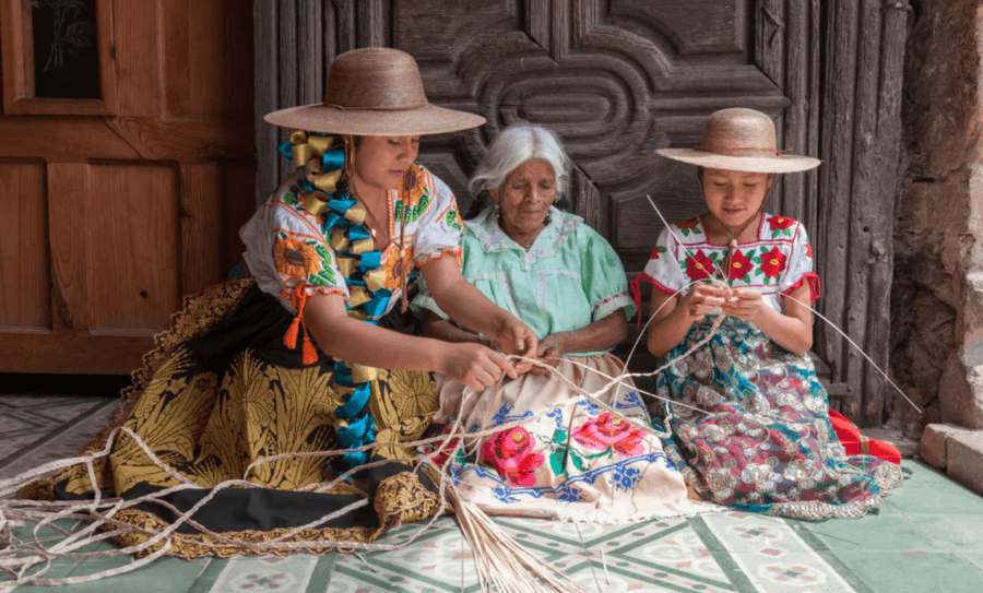Three women in traditional clothing sit on the floor weaving straw for Tula Hats, all in front of a wooden door.