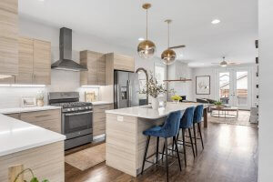 Modern kitchen at 6017B New York Ave, Nashville, featuring light wood cabinets, stainless steel appliances, white countertops, a center island with blue barstools, pendant lights, and an open view to the living area.