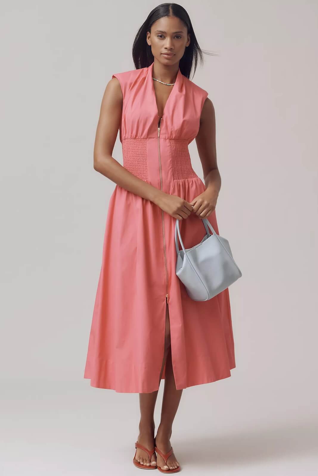 A woman stands wearing a sleeveless coral midi dress with a front zipper and smocked waist, holding a light gray handbag and wearing sandals—it's the kind of dress you'll want one in every color.