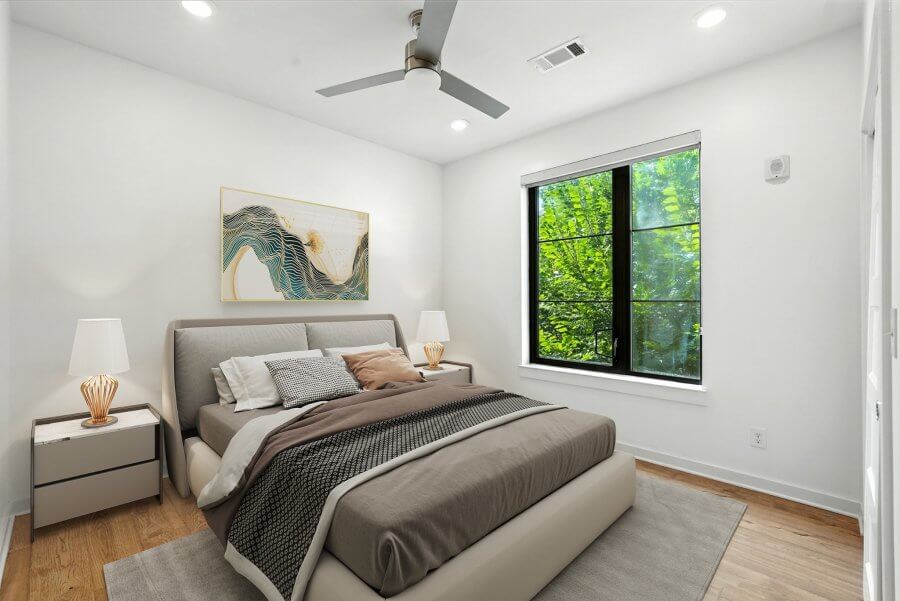 A modern bedroom at 1402 Pillow St, Nashville, featuring a large bed, two nightstands with lamps, a ceiling fan, stylish wall art, and a window revealing lush green trees outside.