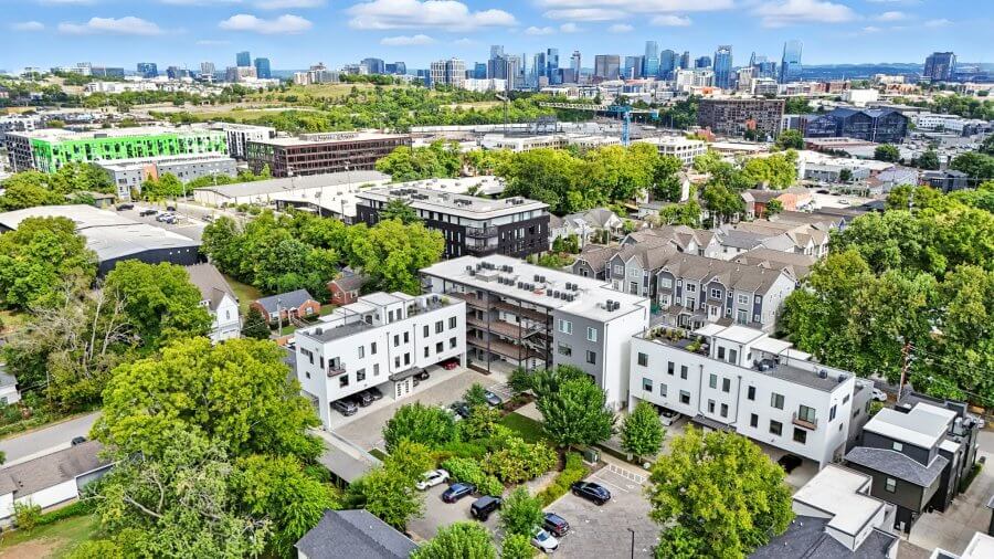 Aerial view of a modern Nashville residential neighborhood near 1402 Pillow St, featuring apartments, townhomes, trees, parking spaces, and a distant city skyline under a partly cloudy sky.