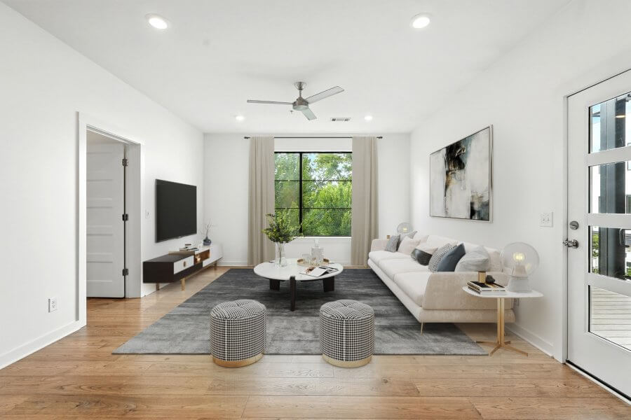 Modern living room on Pillow St with a white sofa, round coffee table, two patterned ottomans, TV on a stand, large window showcasing greenery, and light wood flooring—discover comfort at 1402 Pillow St in Nashville.