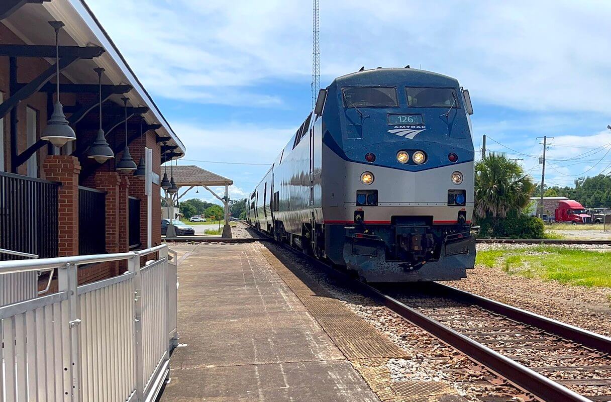 A blue and silver Amtrak passenger train, reminiscent of the festive Amtrak Mardi Gras livery, arrives at a small outdoor station platform on a sunny day.