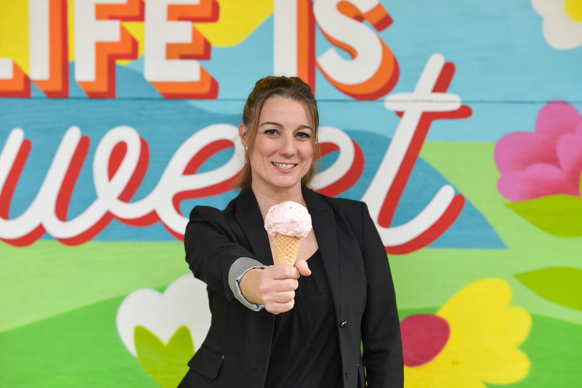 Woman in a black blazer holds out an Andia's Ice Cream cone, standing in front of a colorful mural that says "LIFE IS sweet" with flowers and clouds.