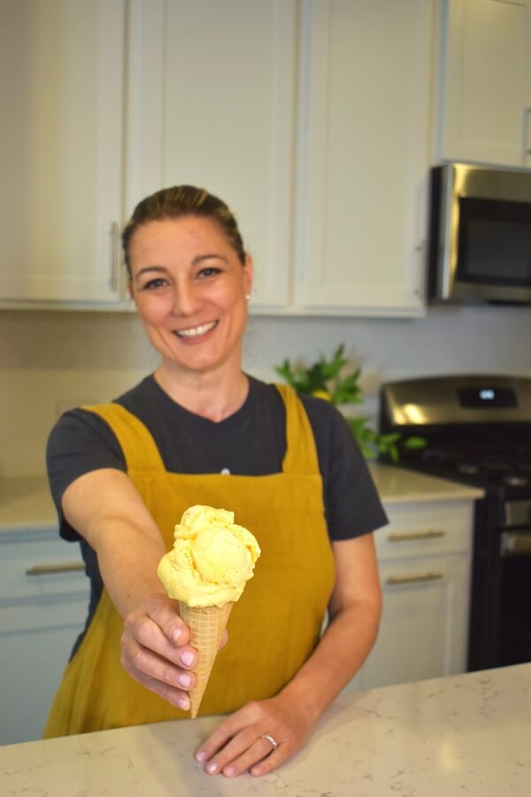 North Carolina's Hottest Ice Cream: Meet Andia's Ice Cream - 6 A woman stands in a kitchen, smiling and holding out an ice cream cone with two scoops of yellow Andia's Ice Cream.