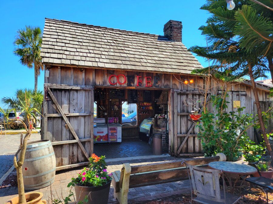 Dreamy coffee shops come to life here—a wooden space with open barn doors, outdoor seating, lush plants, and a bold red "COFFEE" sign above the entrance, all glowing on a sunny day.