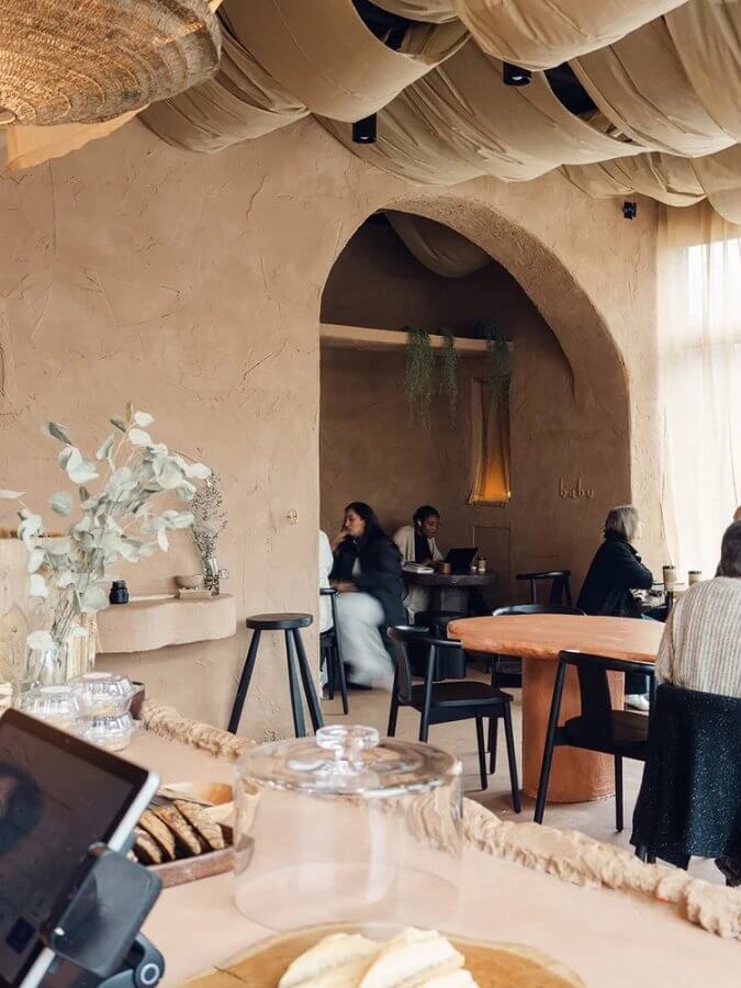 A cozy café with textured beige walls, a draped fabric ceiling, wooden furniture, and several people seated at tables—some eating and chatting—captures the inviting atmosphere of dreamy coffee shops.