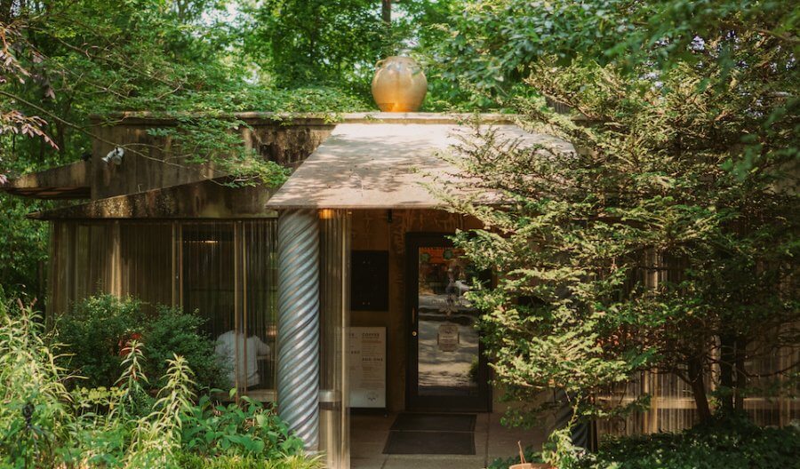 A small building with large windows, a twisted column, and a clay pot on the roof, surrounded by green trees and plants — a dreamy coffee shop tucked away in nature.