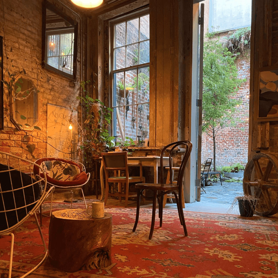 A cozy room with eclectic furniture, wooden walls, a red patterned rug, potted plants, and an open door leading to a brick courtyard with greenery.