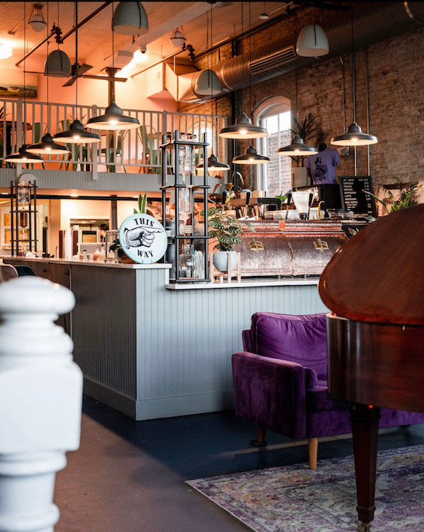 A cozy café interior reminiscent of dreamy coffee shops, with exposed brick walls, a mezzanine, industrial lighting, a counter marked by a "This Way" sign, and plush purple velvet seating.