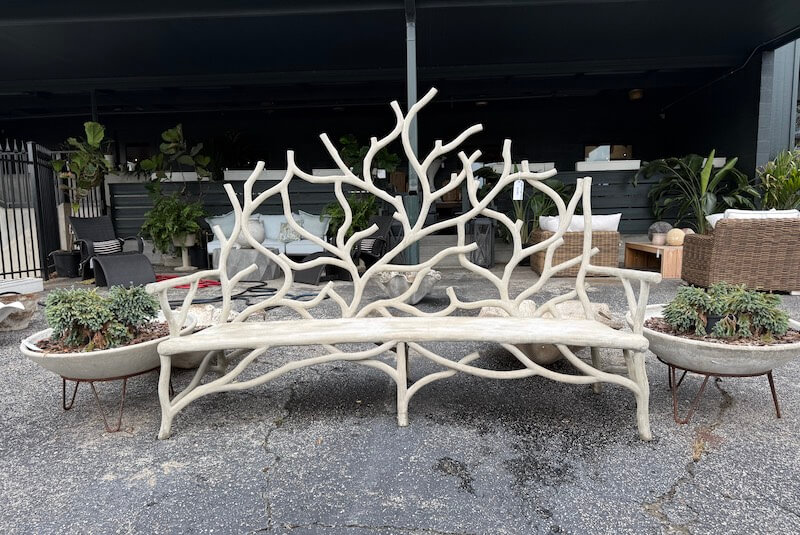 A sculptural outdoor bench with a tree-branch design stands between two large planters with succulents, a highlight among southern finds August collections, positioned on a paved surface in front of patio furniture.