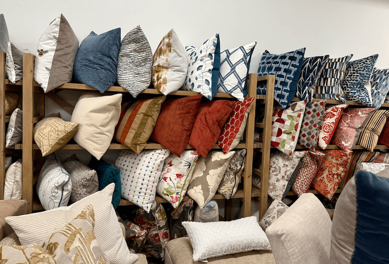 Shelves filled with assorted decorative pillows in various colors, patterns, and textures, displayed against a white wall at Margi’s Furniture—where exceptional furniture design meets style in Green Hills.