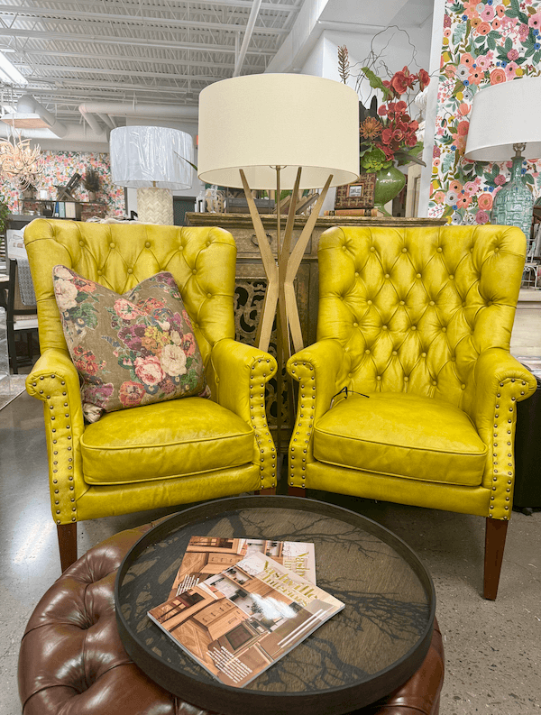 Two yellow tufted armchairs from Margi’s Furniture sit side by side with a floral pillow on one, a round coffee table with magazines in front, and a tripod floor lamp behind them in the Green Hills showroom.
