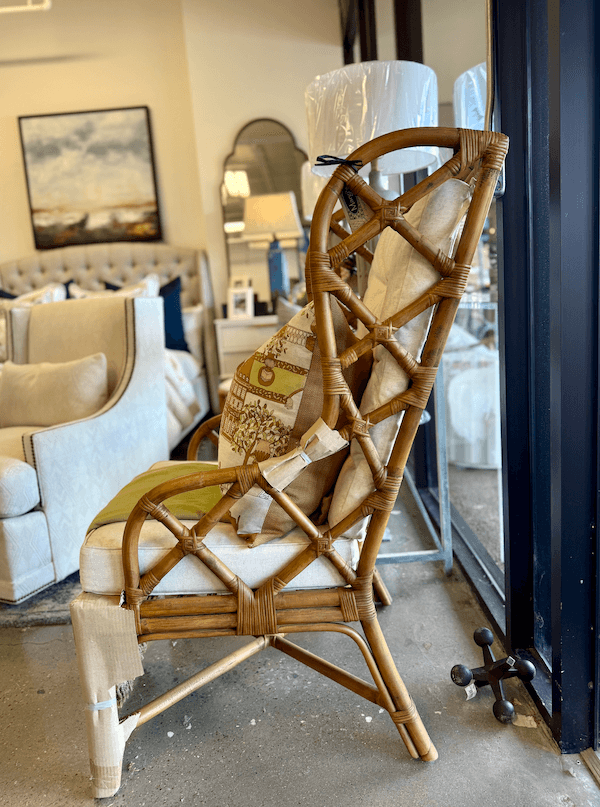 A rattan armchair with a cushion sits by a large window at Margi’s Furniture, surrounded by other upholstered chairs and decorative items, showcasing elegant furniture design in Green Hills.