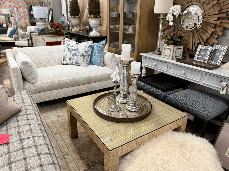 A living room showroom at Margi’s Furniture in Green Hills displays a white patterned sofa, glass-top coffee table with candles, a desk, framed photos, and assorted decor that highlight elegant furniture design.