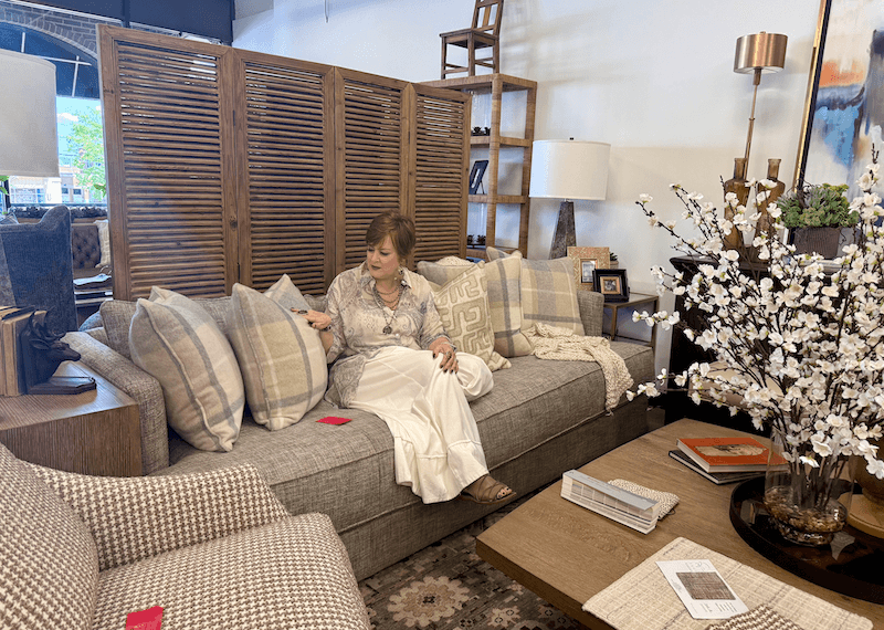 A woman sits on a gray sofa with multiple pillows in a modern living room showroom at Margi’s Furniture, featuring stylish wood furniture and decorative accents inspired by contemporary Furniture Design.