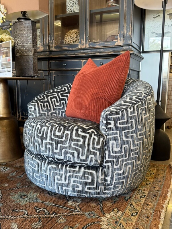 A patterned black and white swivel chair from Margi’s Furniture, accented with a textured rust-colored pillow, sits on an ornate rug in front of a dark cabinet—showcasing exceptional furniture design.