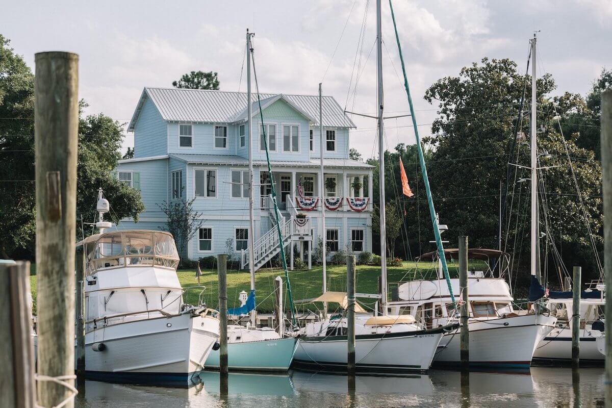 Several boats are docked in front of a light blue, two-story house decorated with patriotic bunting in Ocean Springs, MS, surrounded by trees and grass on a sunny day.