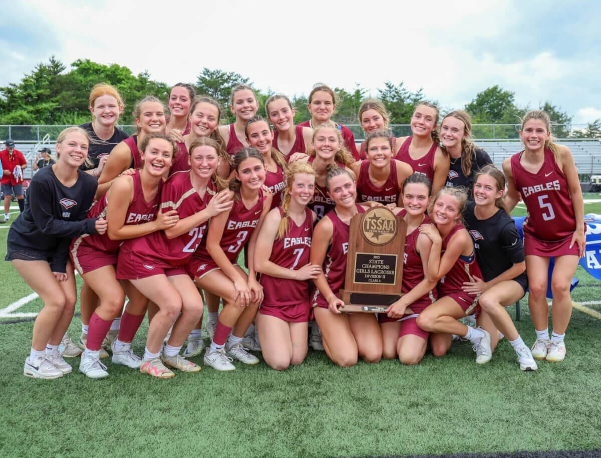 A girls’ lacrosse team in maroon uniforms poses together on a field, smiling and holding a large championship trophy.