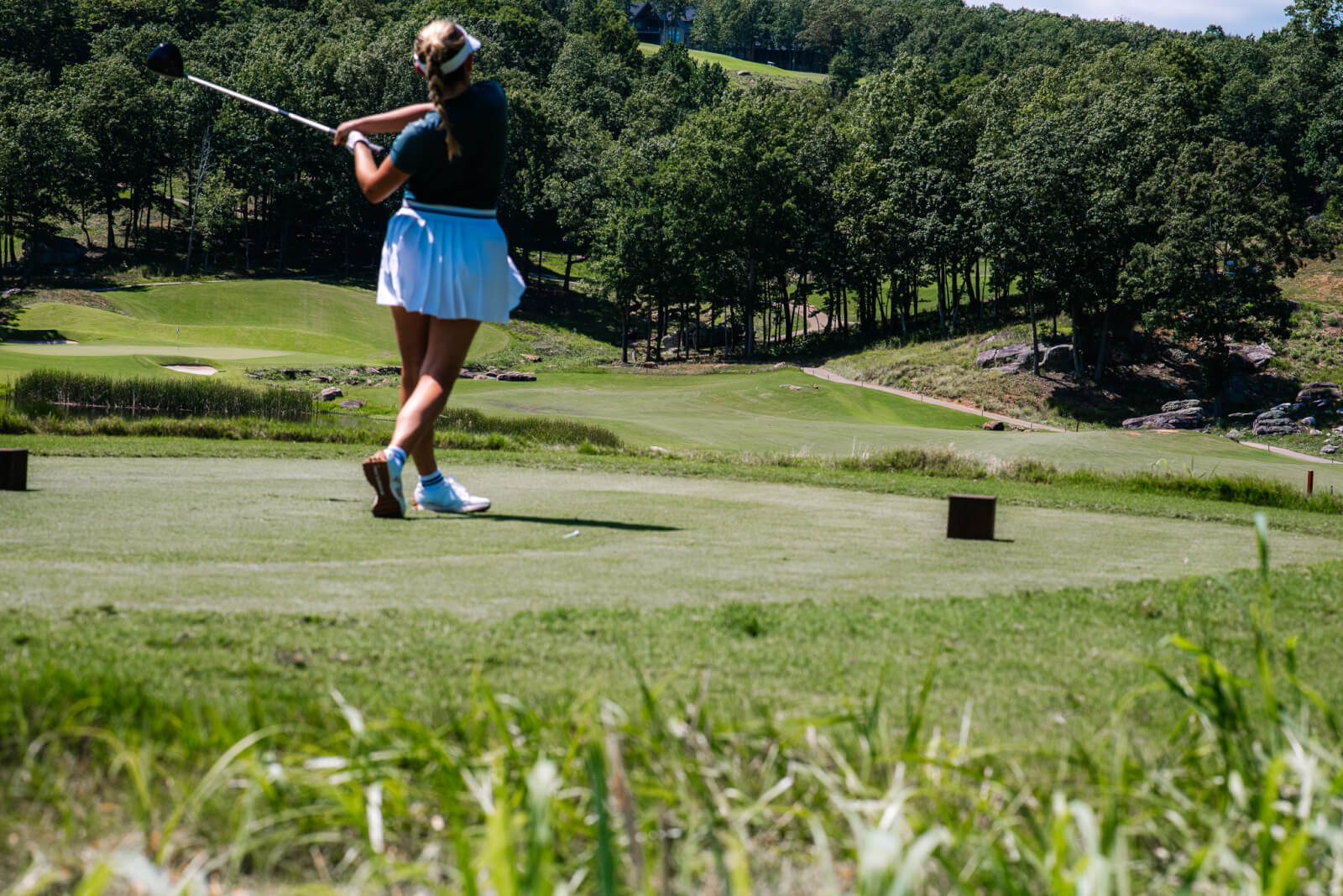 A golfer wearing a white skirt and black top follows through on a swing at a tee box at McLemore Resort, with a grassy golf course and trees in the background.