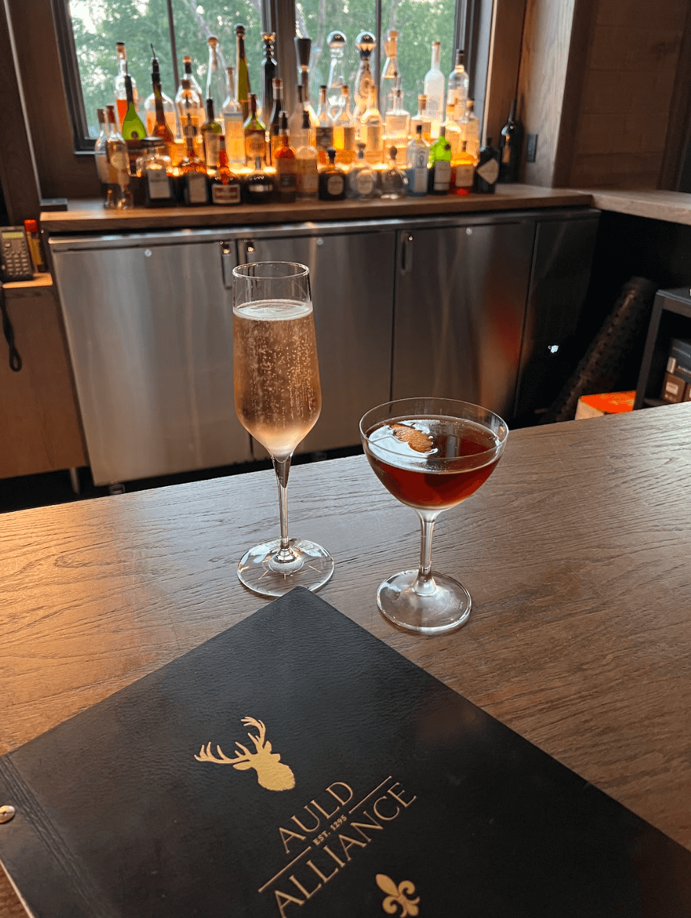 Two cocktails sit on a bar counter beside an Auld Alliance menu at the elegant McLemore Resort, with a selection of liquor bottles displayed on shelves in the background.