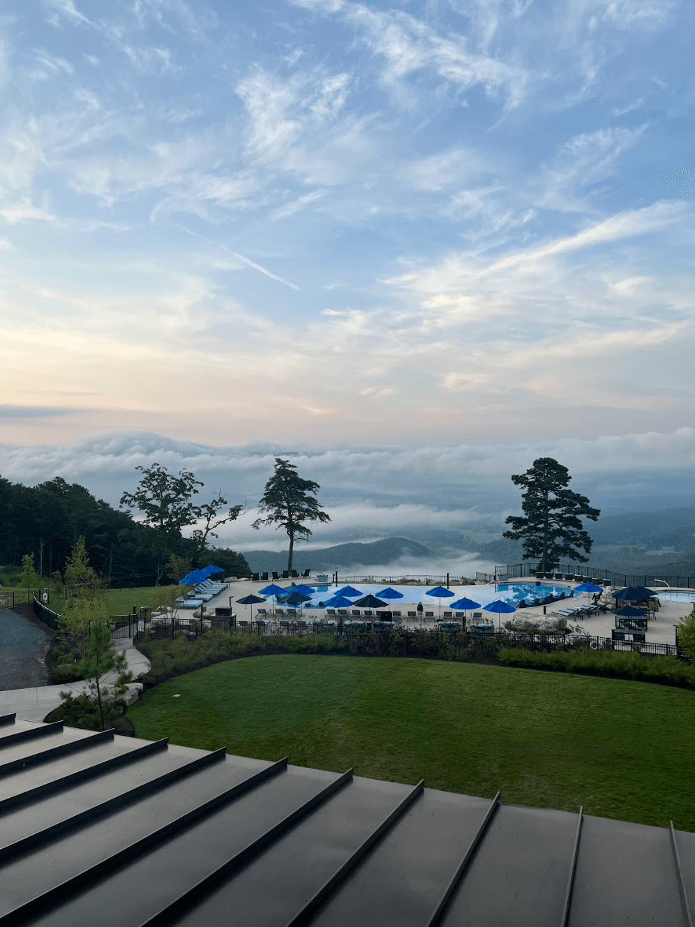 Outdoor swimming pool area with blue umbrellas at McLemore Resort overlooks a misty valley and distant hills under a partly cloudy sky at sunrise or sunset.