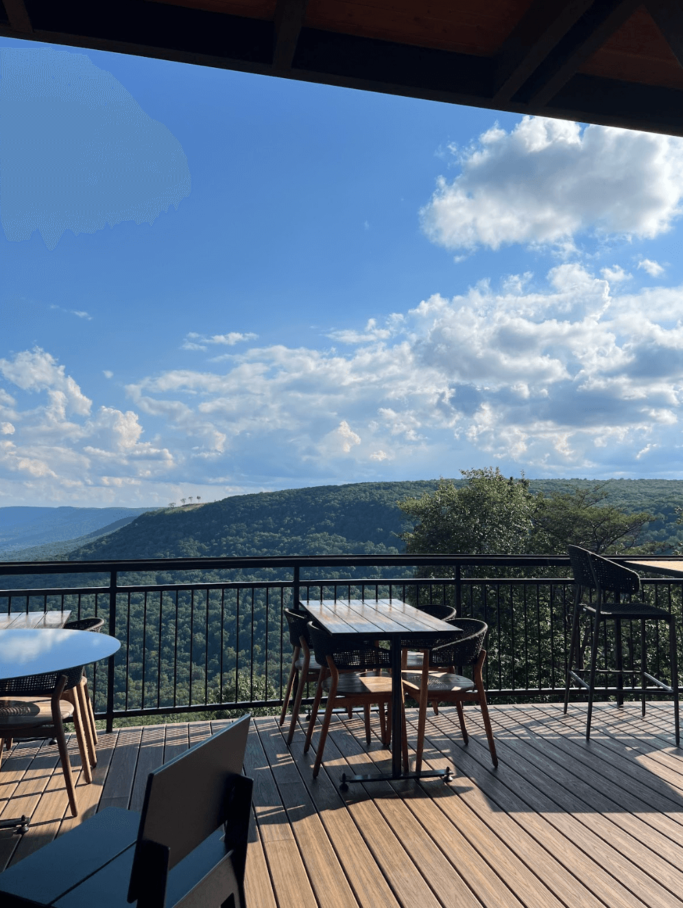 Outdoor dining area with empty tables and chairs on a wooden deck at McLemore Resort overlooks a green, tree-covered hill under a blue sky with scattered clouds.