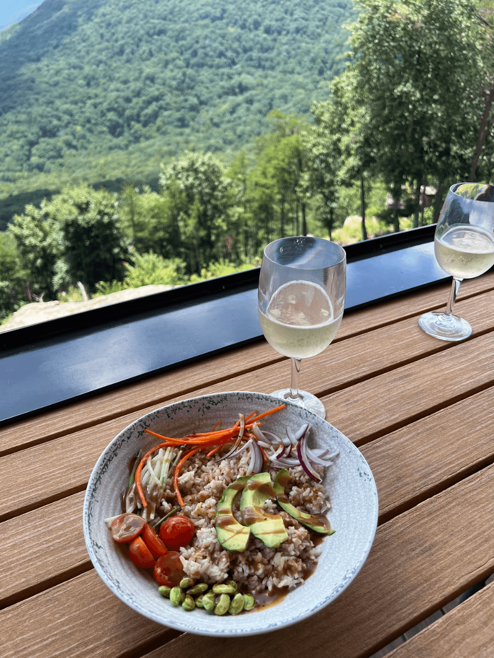 A bowl of rice salad with vegetables and avocado on a wooden table, two glasses of white wine, and a stunning green mountain view at McLemore Resort in the background.