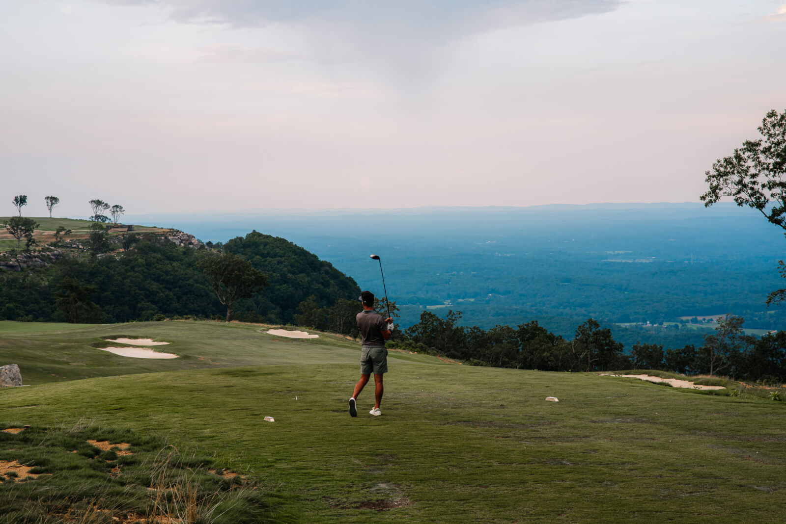 A golfer swings a club on the lush green course at McLemore Resort, with sand bunkers nearby and hills overlooking a wide, forested valley under a cloudy sky.
