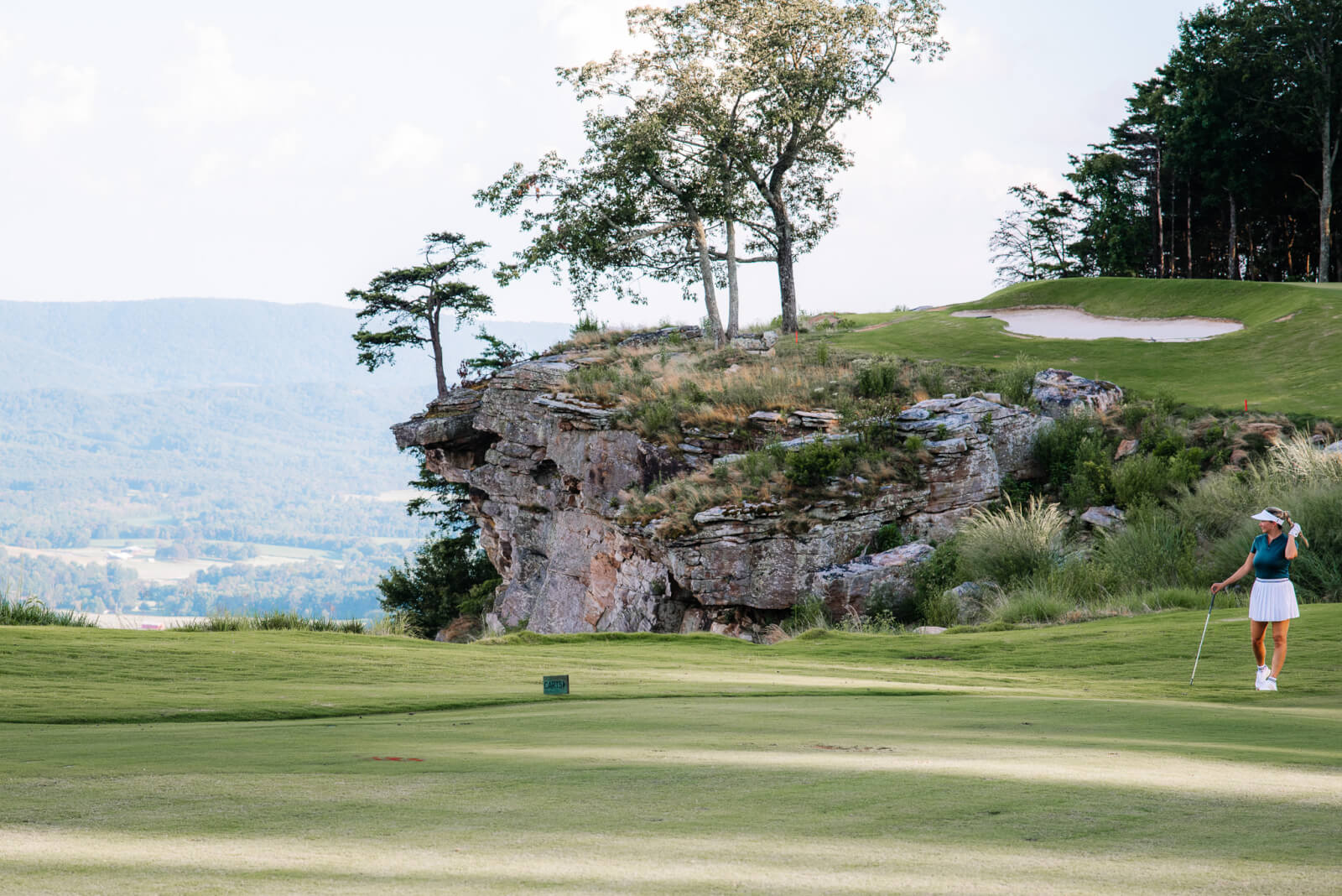 A golfer stands on a green fairway at McLemore Resort beside a rocky cliff, with trees and mountains visible in the background.