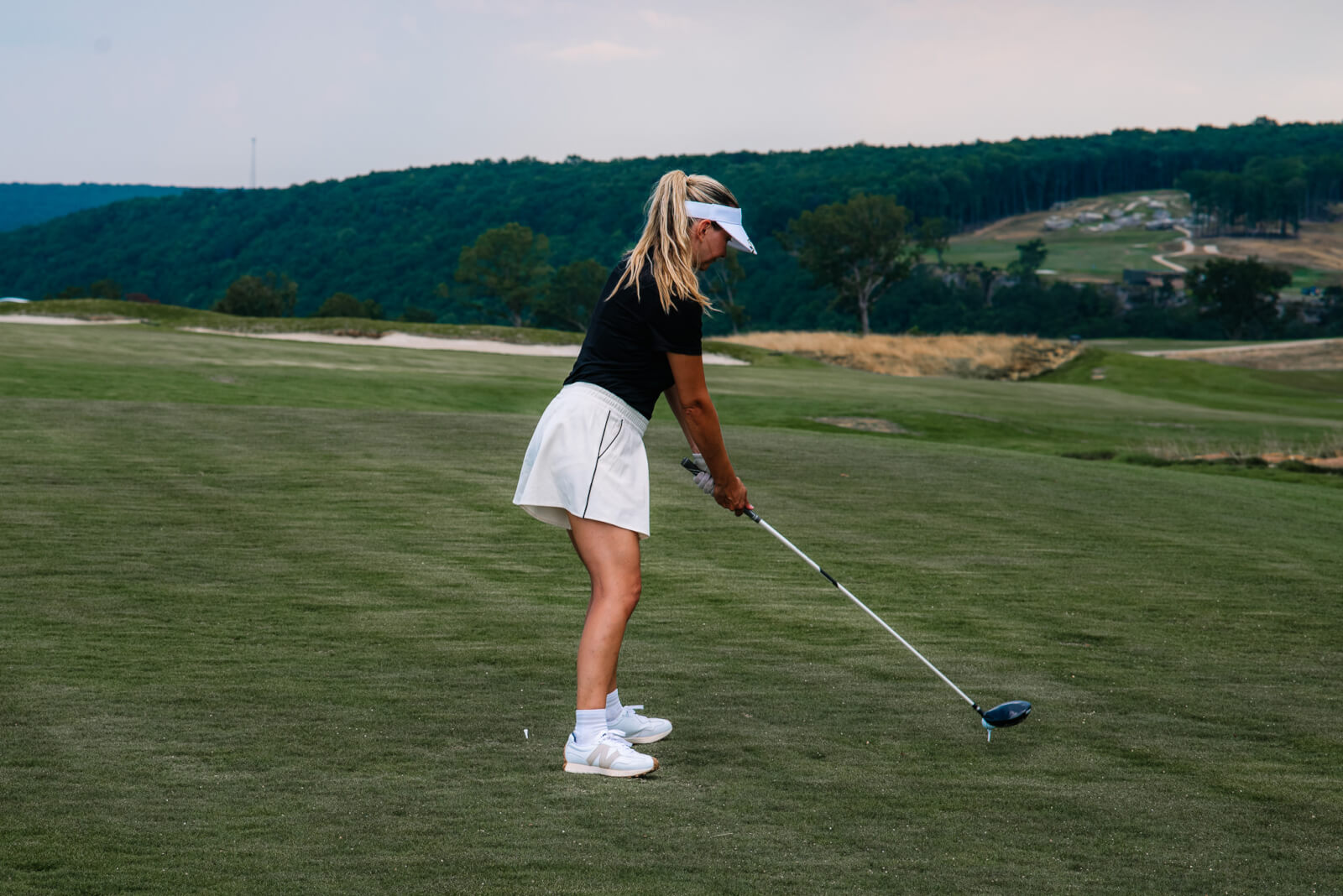 A woman in athletic attire prepares to hit a golf ball with a club on the lush, rolling greens of McLemore Resort, with scenic hills and trees lining the background.