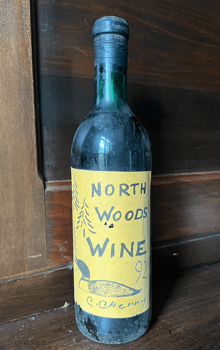 A dusty bottle labeled "North Woods Wine 92 C. Cherry" with hand-drawn trees and a bird evokes the charm of lake life, set against a rustic wooden background.