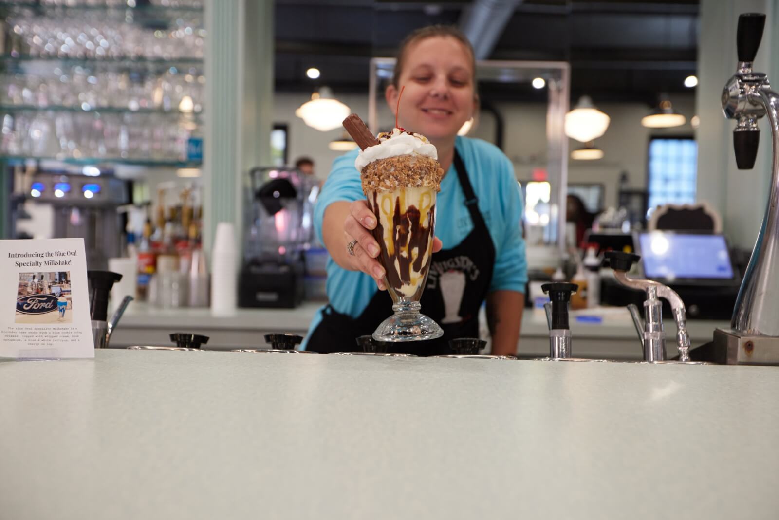 Culinary Road Trip: 4 Tennessee Towns for Food Lovers - 2 A server behind a counter presents a tall sundae glass filled with ice cream, whipped cream, caramel sauce, and a chocolate stick—a must-try treat for food lovers on a Tennessee culinary road trip.