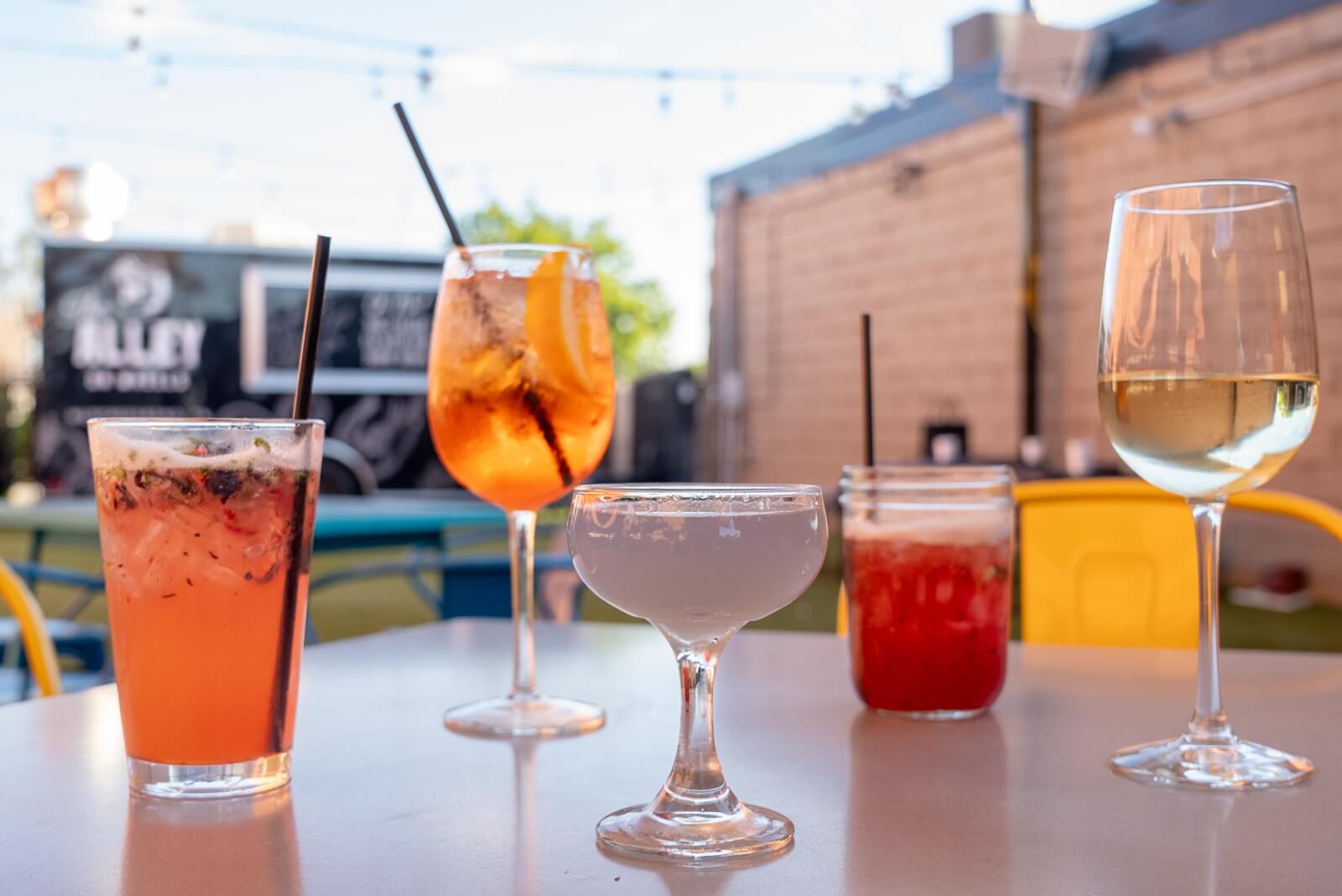 Culinary Road Trip: 4 Tennessee Towns for Food Lovers - 6 Five assorted cocktails in different glasses with straws sit on an outdoor table, perfect for food lovers enjoying a culinary road trip, with blurred chairs and a food truck visible in the background.