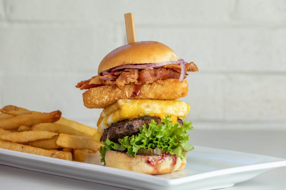 A tall burger stacked with lettuce, tomato, cheese, hamburger patty, onion rings, bacon, and onion on a bun with French fries, showcasing the bold flavors found in new restaurants and dining concepts across Birmingham.