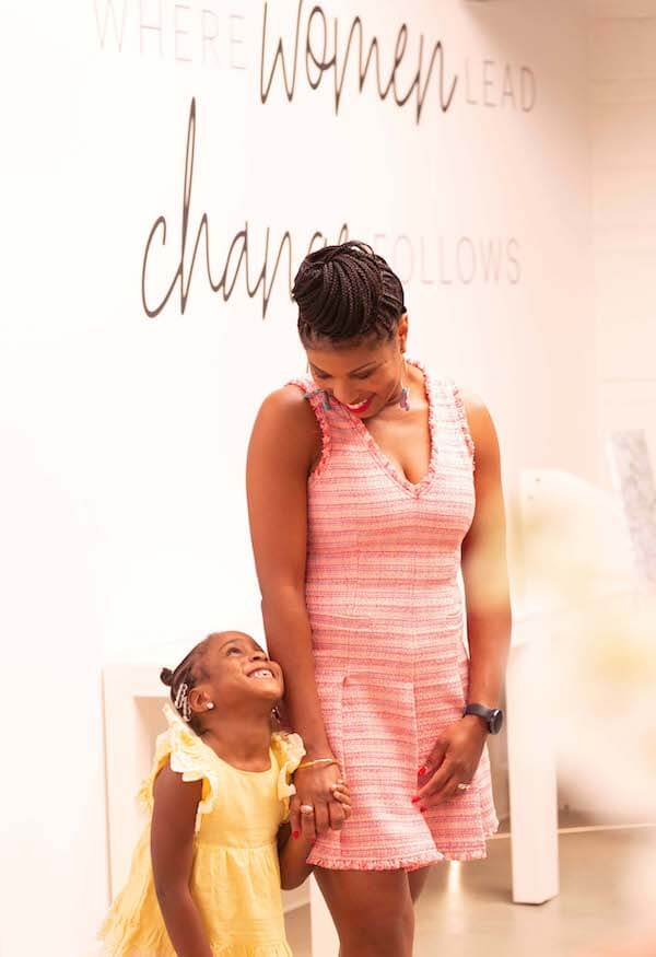 A woman in a pink dress, the new CEO of Women's Foundation of Alabama, holds hands with a smiling young girl in a yellow dress. The wall behind them reads, "Where women lead, change follows.