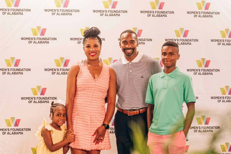 A family of four stands in front of a Women’s Foundation of Alabama step-and-repeat banner, smiling for the camera and celebrating the arrival of the new CEO.