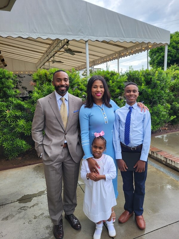 A family of four poses outdoors; the father, the new CEO of the Women's Foundation of Alabama, wears a suit, the mother a blue dress, the son a shirt and tie, and the daughter a white dress with a pink bow.